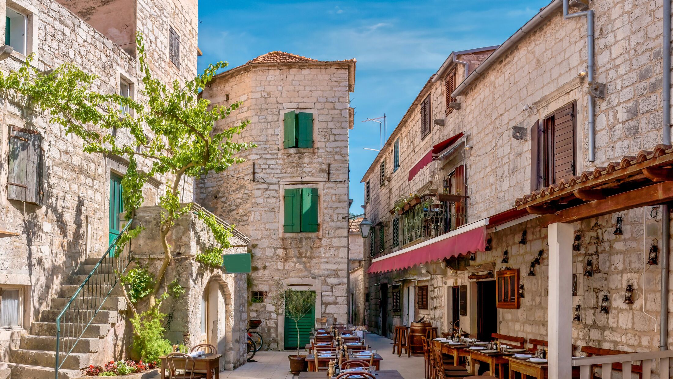 Street view of a charming outdoor tourist restaurant, open and prepared for customers, with rustic wooden furniture and surrounded by old stone buildings, in Stari Grad, Hvar Island, Croatia.