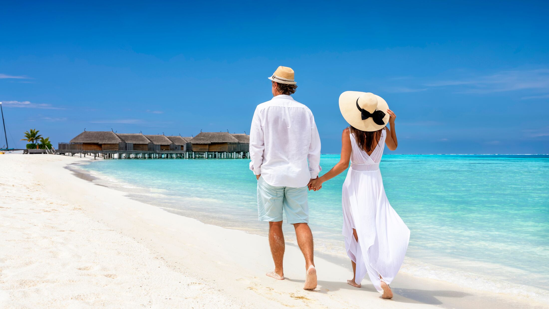 Happy couple in white clothing and with hats walks down a tropical beach with turquoise sea in the Maldives islands