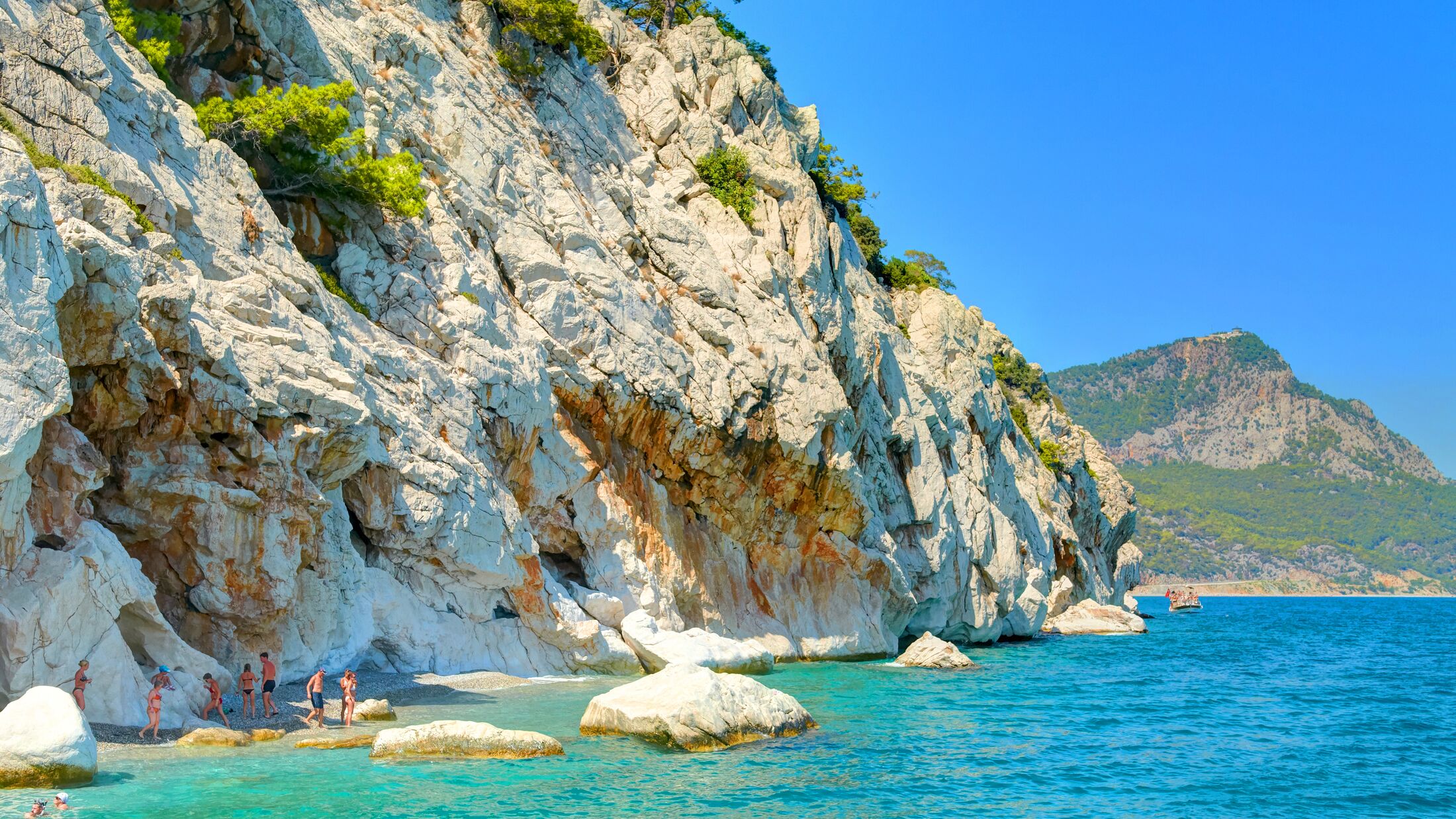 Sand color stone rocks beach with green trees in the Mediterranean Sea Turkey seaside. Swimming people tourists. Best Turkey holidays vacations famous boat tours on Mediterranean Sea. Romantic beach